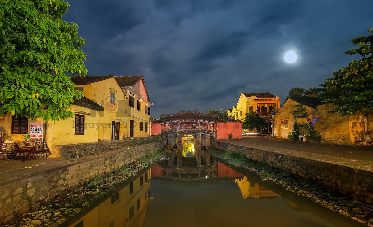 The Japanese Covered Bridge in Hoi An lit up at night under a full moon, reflected in the canal below