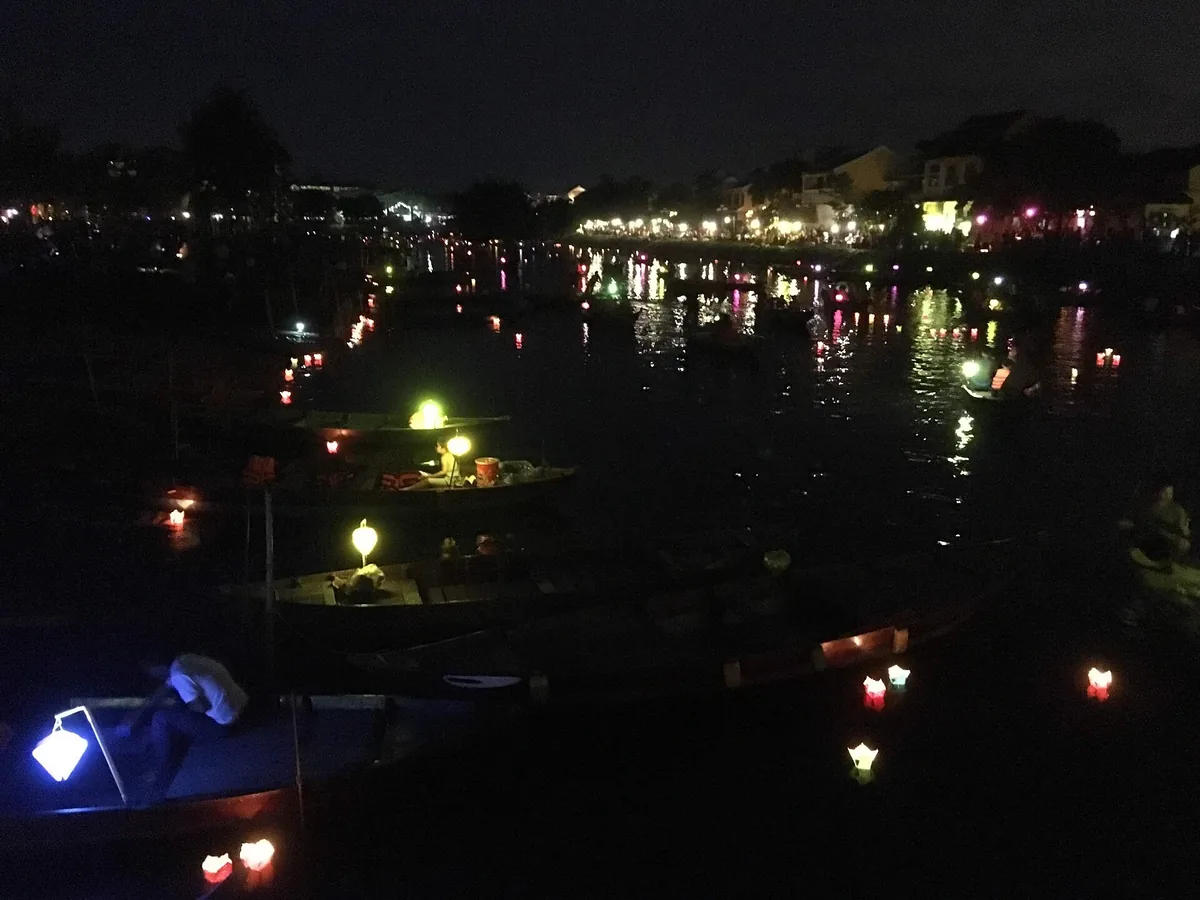 Small lanterns floating on the dark Thu Bon River at Hoi An during the full moon festival, with lit houses along the far bank