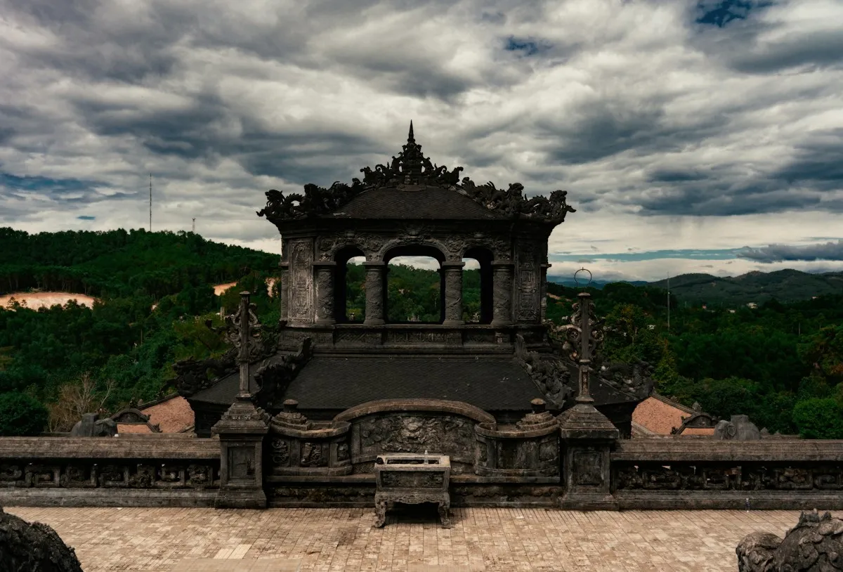 The dark stone stele pavilion at the tomb of Emperor Khai Dinh under heavy clouds, with forested hills behind