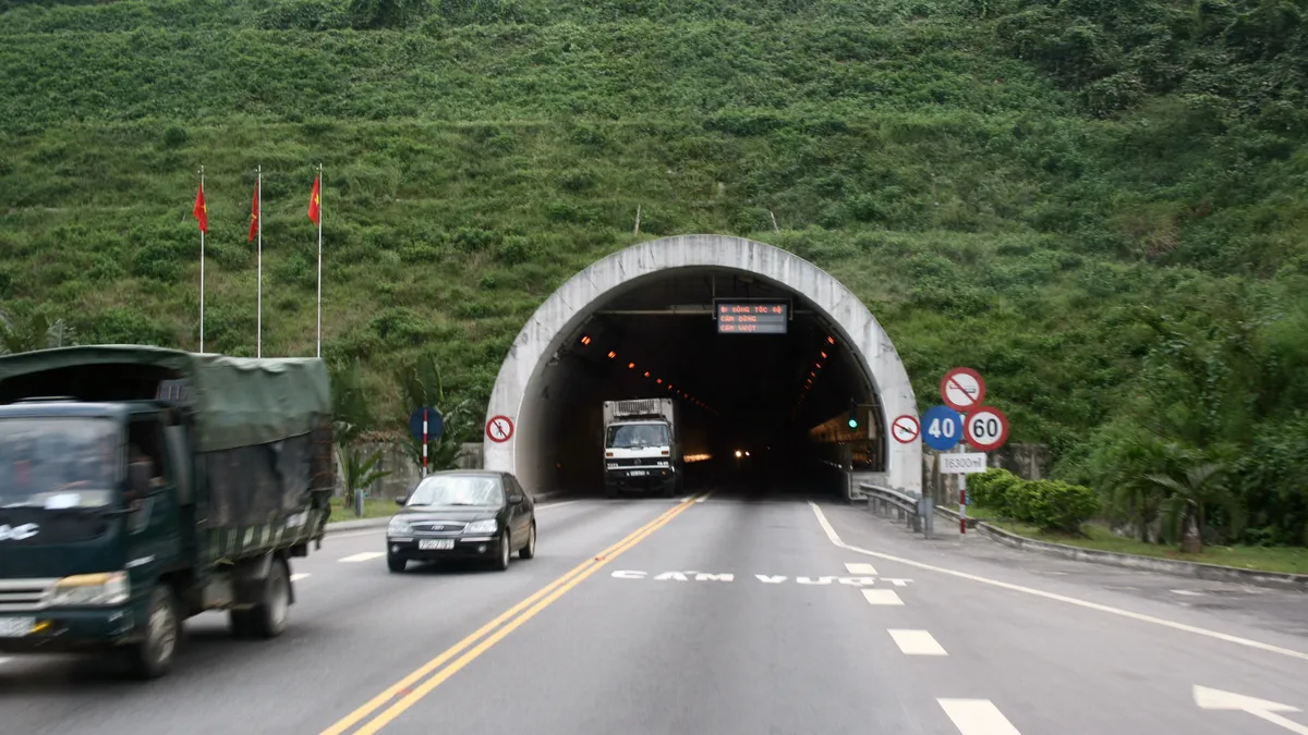 A truck and a car driving out of the arched north entrance of the Hai Van road tunnel, green hillside above