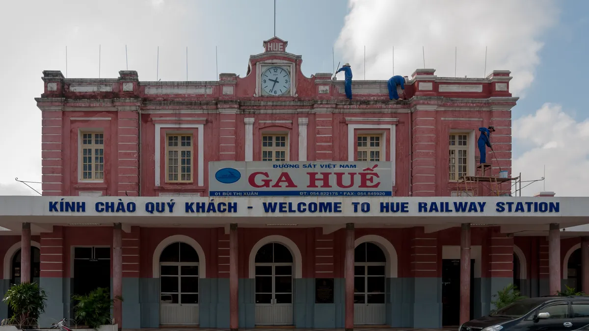 The pink colonial-style facade of Hue railway station with its clock and a blue Ga Hue sign, workers painting the upper floor