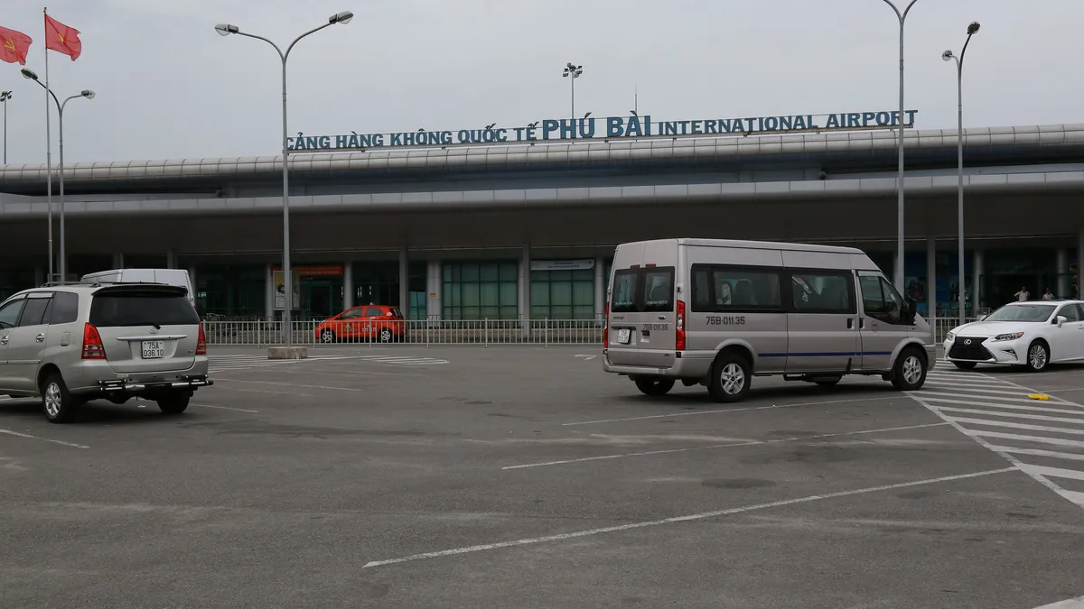 The front of Phu Bai airport terminal under a grey sky, its name across the roof, a silver van and two cars in the car park