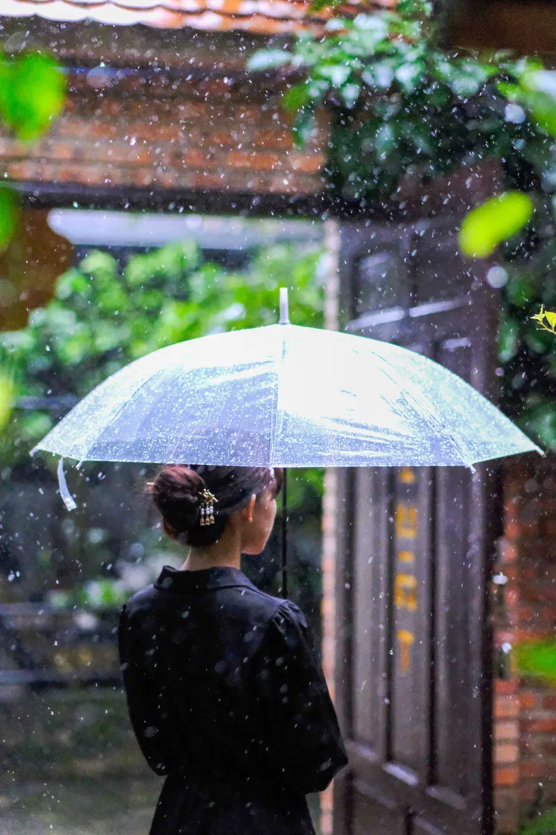 A woman in a black dress holding a clear umbrella in heavy rain beside a brick gateway and wet greenery in Hue