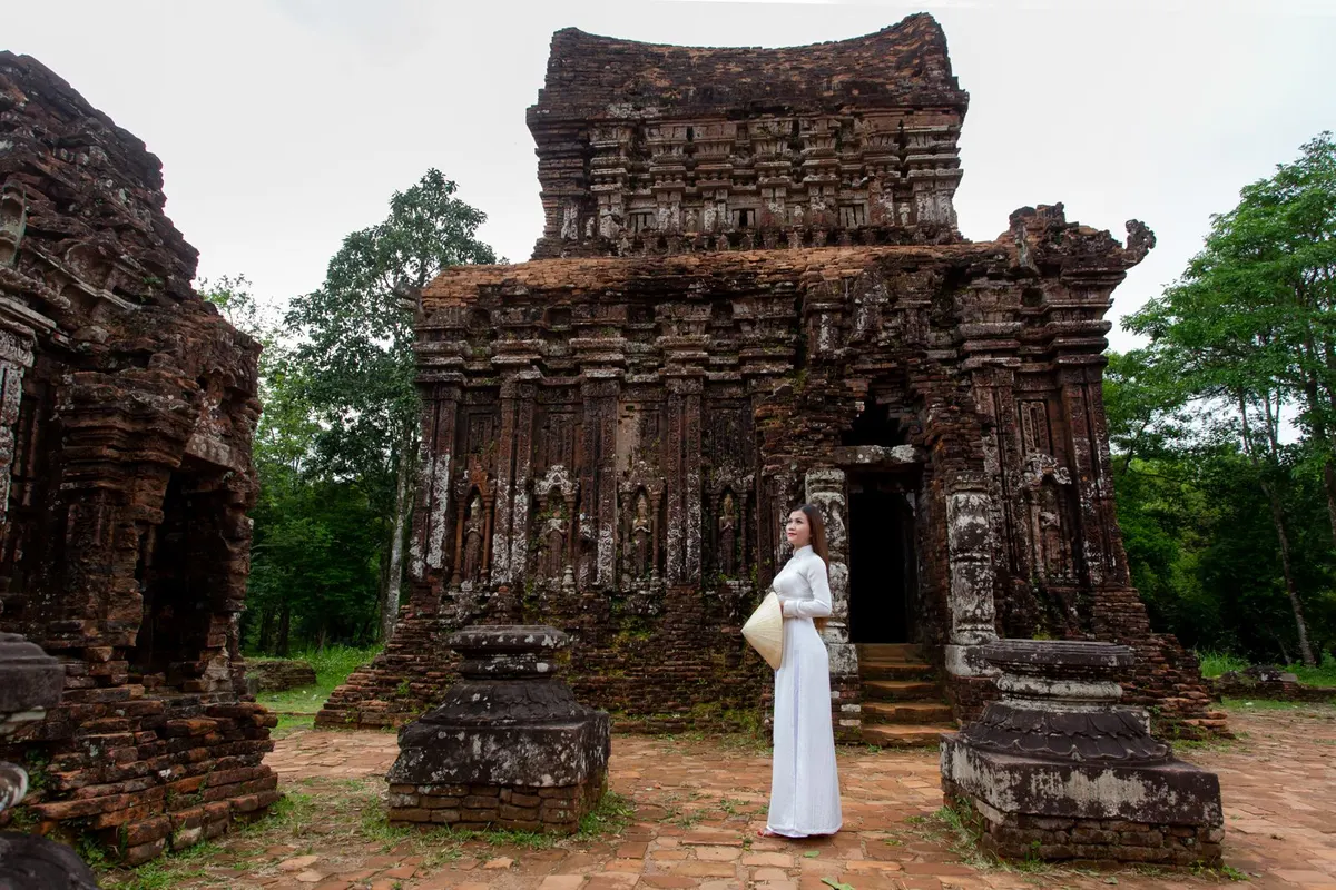 A woman in a white ao dai holding a conical hat stands before a carved brick Cham temple at My Son