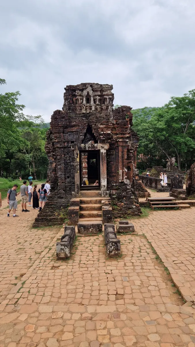 Visitors with backpacks gather beside a ruined brick Cham tower on the paved grounds of My Son Sanctuary