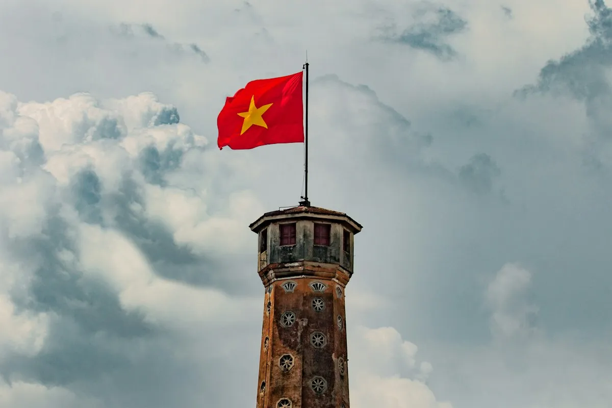 The brick top of the Hanoi Flag Tower with a red Vietnamese flag against grey and white clouds