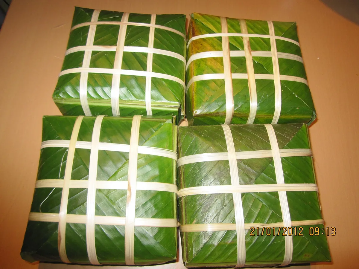 Four square banh chung rice cakes wrapped in green dong leaves and tied with bamboo strips, on a table