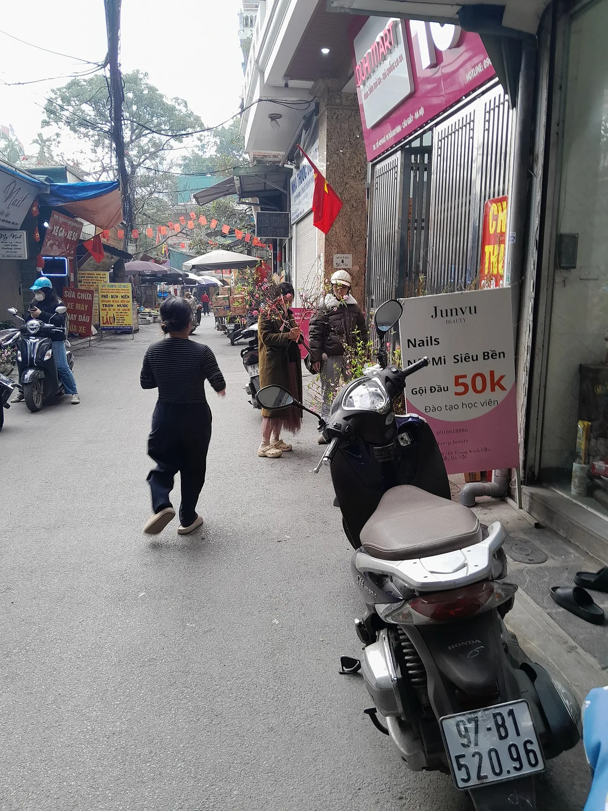 Shoppers choosing peach blossom branches at a street stall in a narrow Hanoi lane hung with red flags and lanterns