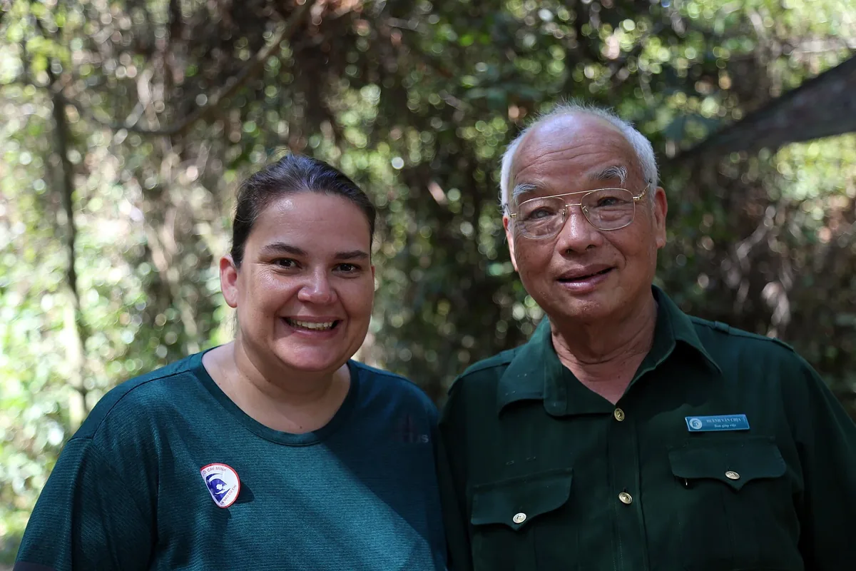 An older man in a green uniform with a name badge standing beside a smiling visitor among trees at the Cu Chi Tunnels
