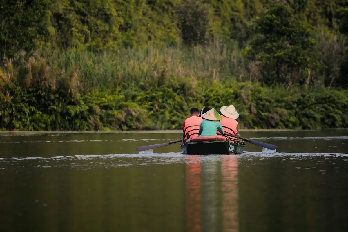 Two visitors in conical hats and orange life jackets rowed along a quiet green-lined river in Trang An