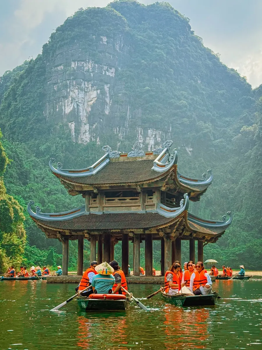 Rowers in conical hats steer sampans of visitors in orange life jackets past a river pavilion below a limestone cliff