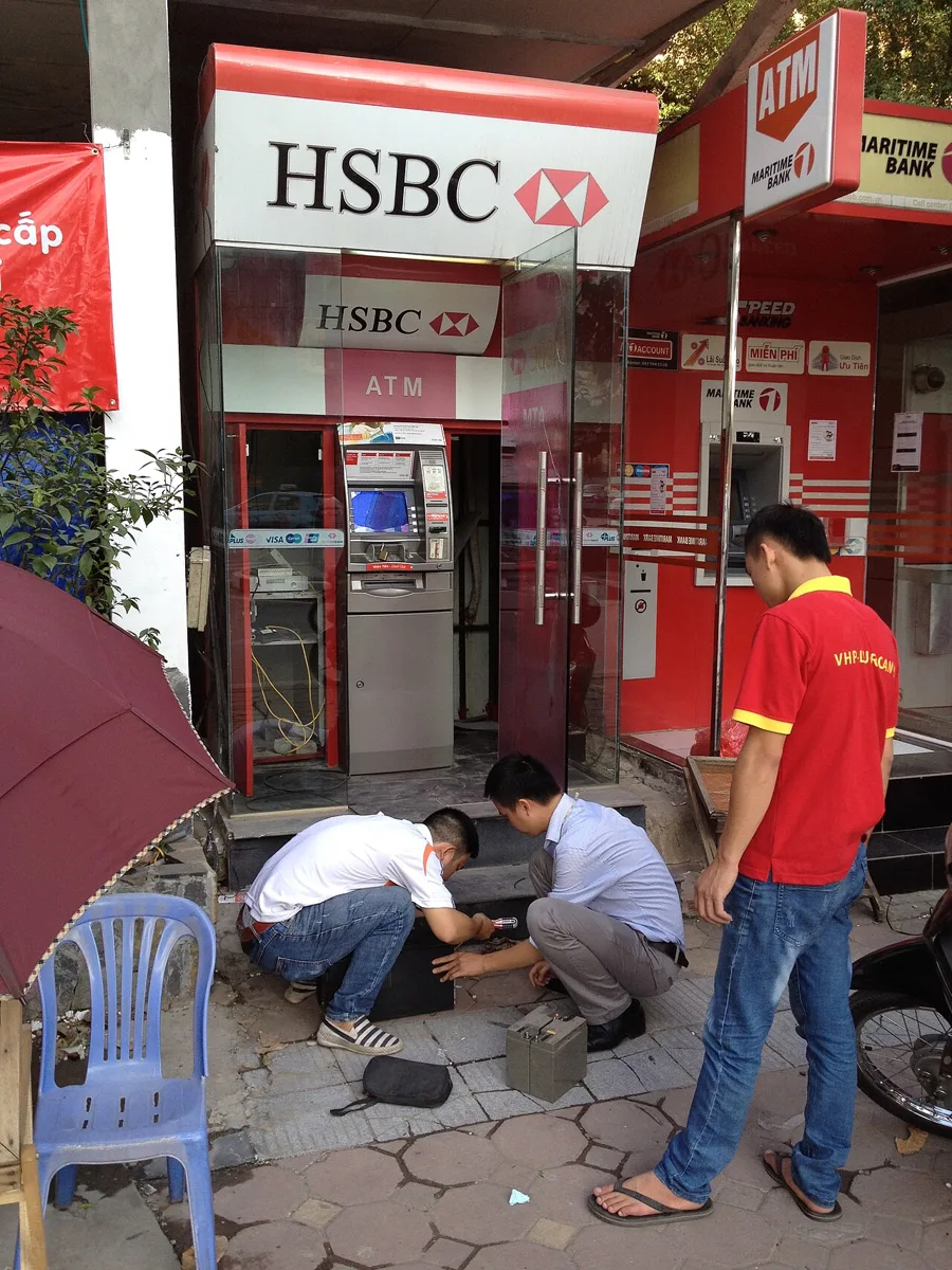An HSBC ATM booth on a Hanoi pavement with two men kneeling at its base, beside a red Maritime Bank ATM booth