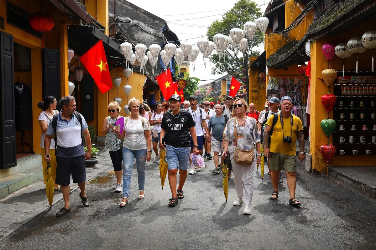 A large group of foreign tourists walking down a lantern-hung street of yellow shophouses with Vietnamese flags overhead