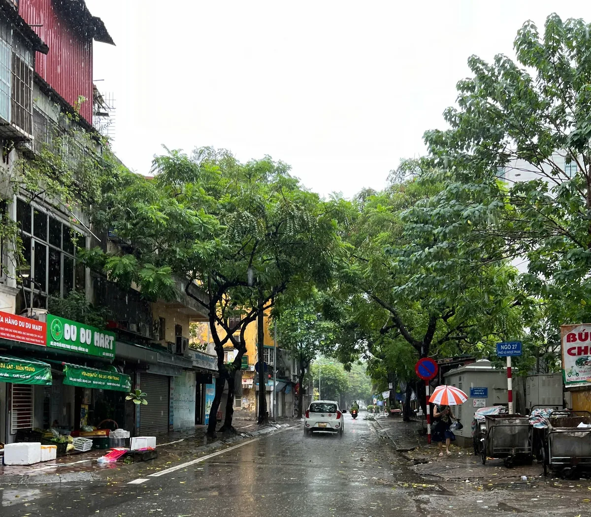 A wet street in Dong Da, Hanoi, with a pedestrian under an umbrella and a car on the road