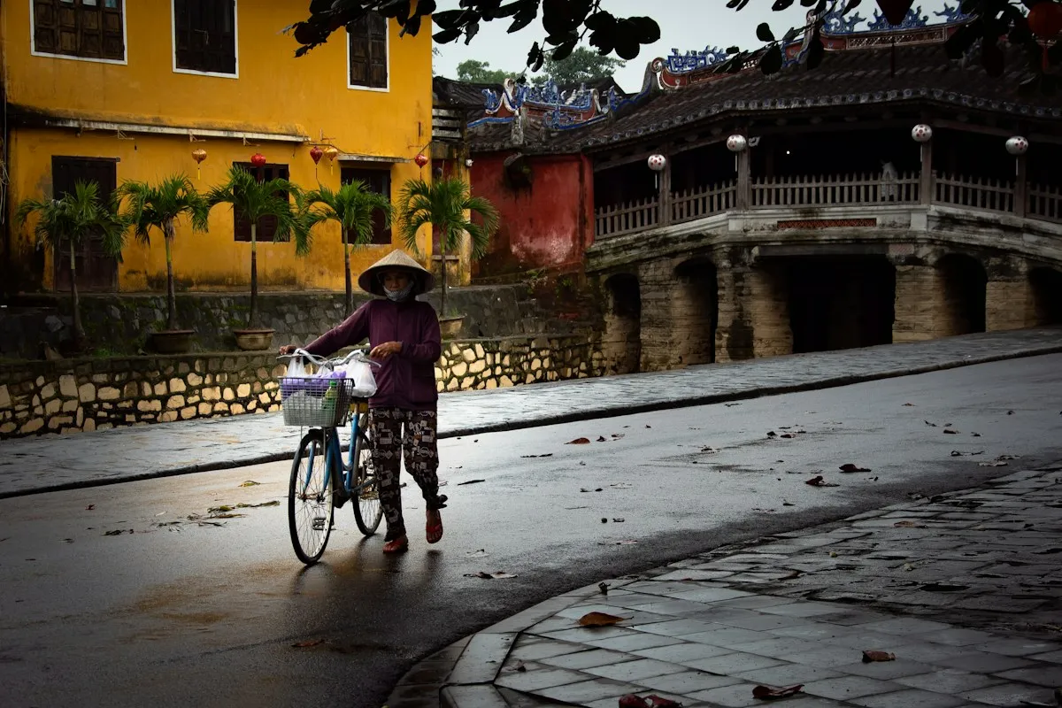 A woman in a conical hat pushing her bicycle along a wet street beside the Japanese Covered Bridge in Hoi An