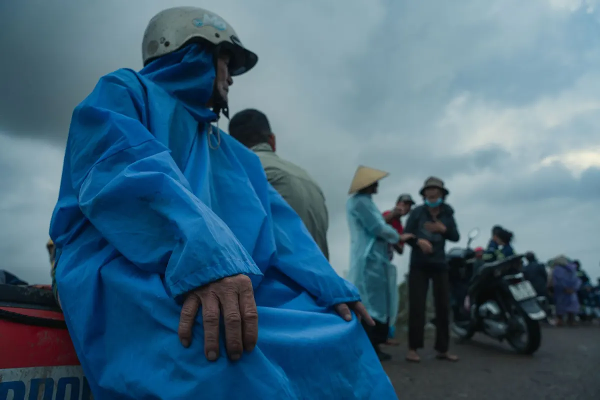 An older man in a blue rain poncho and helmet beside people in raincoats and motorbikes under a dark November sky in Phu Yen