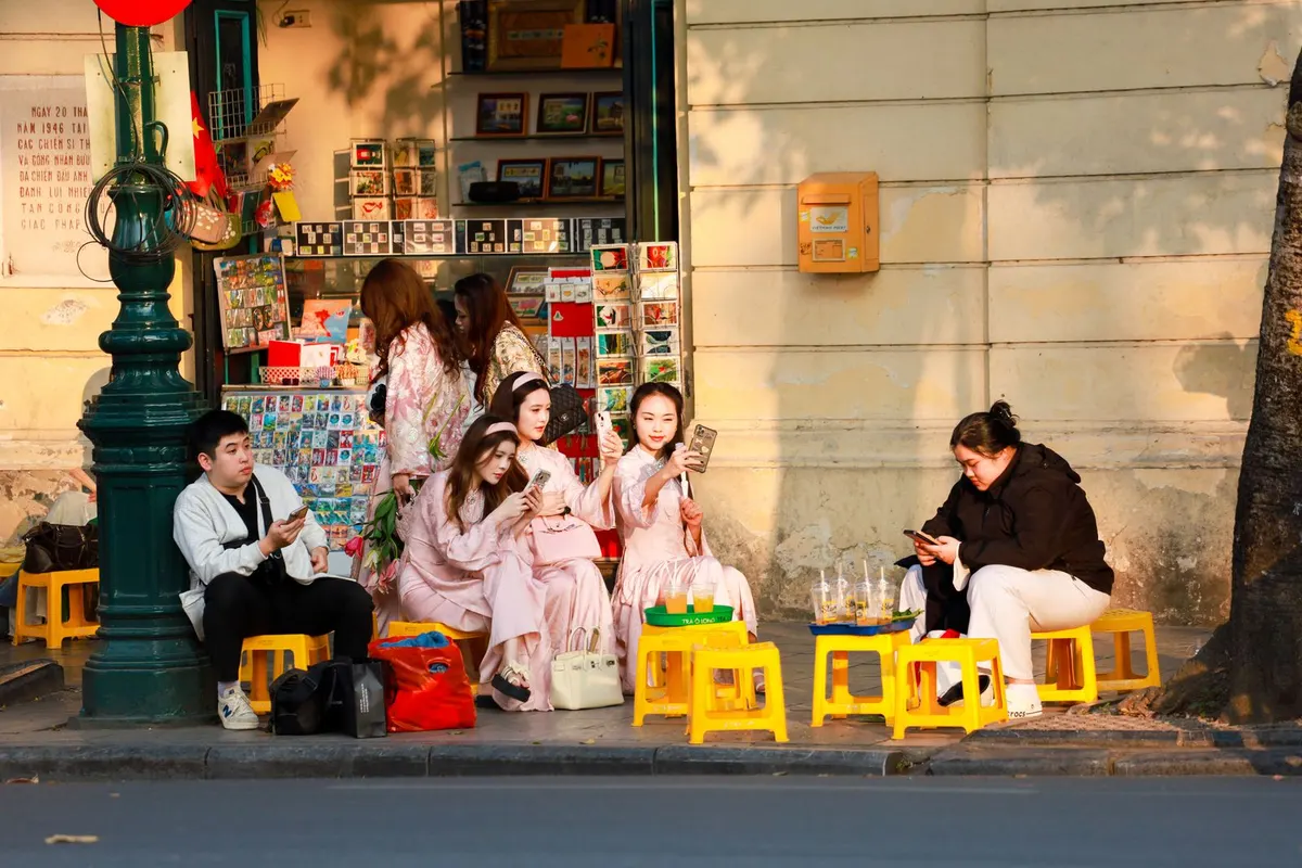 Young people on yellow plastic stools outside a Hanoi postcard shop, most of them looking at their smartphones