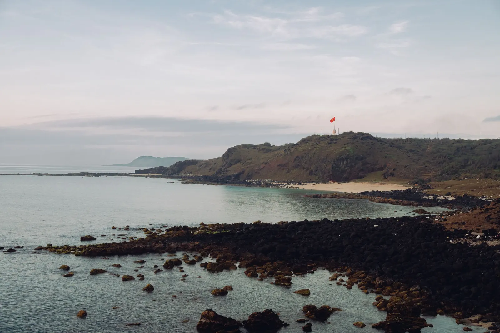 The flag on a headland above a sandy cove and black volcanic rocks on Phu Quy