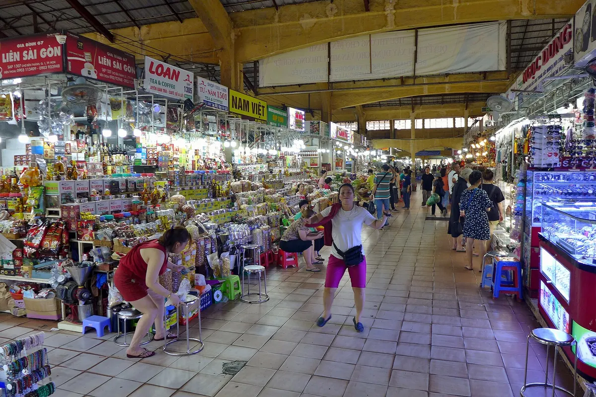 A long aisle under a yellow steel roof, lined with stalls selling snacks and souvenirs, shoppers walking through