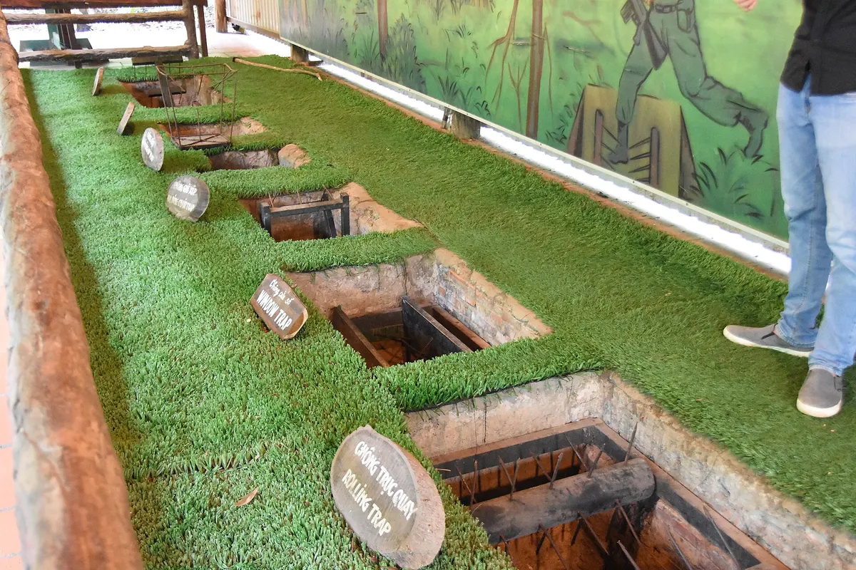 A row of booby-trap pits with wooden spikes set into fake grass, each with a small name sign