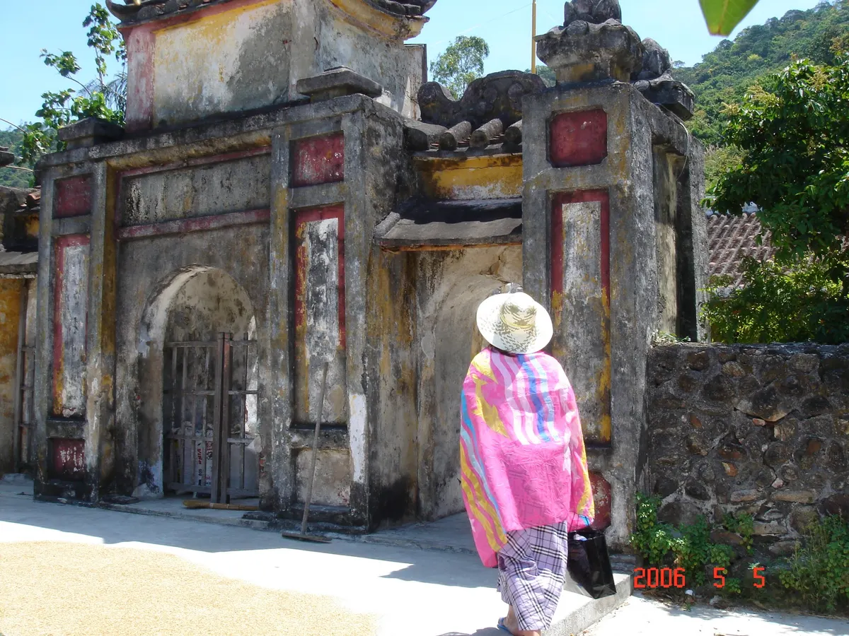 An old pagoda gate with a curved roof and faded red and yellow walls, with a woman in a conical hat walking past
