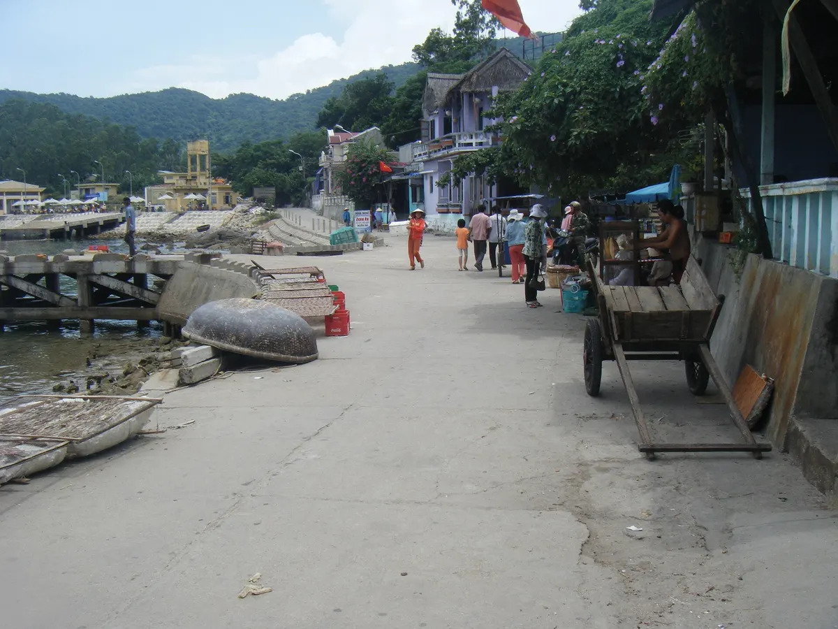 A waterfront street on Cu Lao Cham with houses and a handcart on one side, round basket boats on the rocky shore and green hills behind
