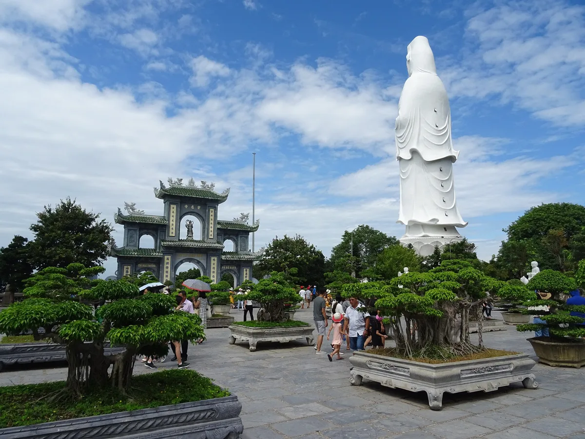 A tall white statue of the Lady Buddha above a paved courtyard with bonsai trees and an ornate gate at Linh Ung Pagoda