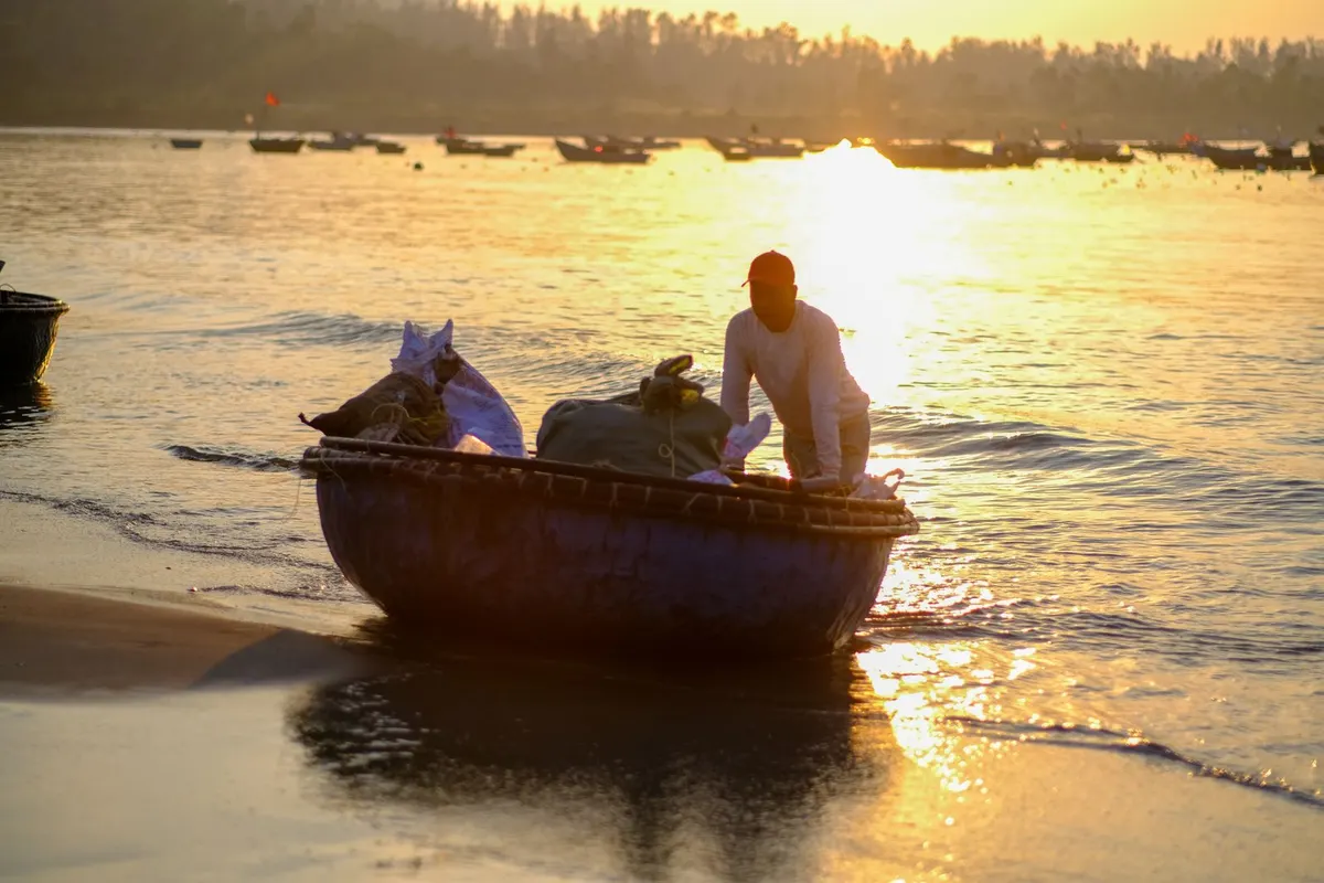 A fisherman in a red cap pushes a loaded round basket boat onto the sand at sunrise, more boats moored behind