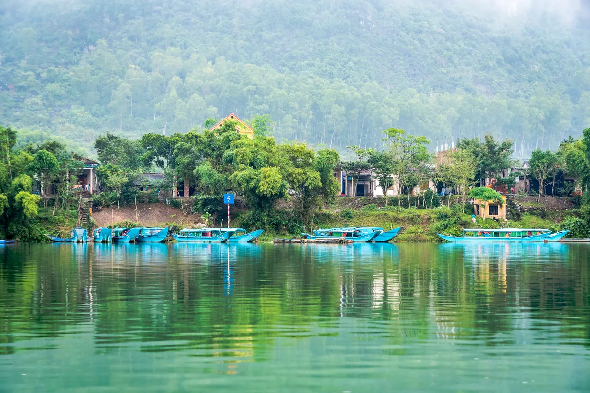 Turquoise boats moored on the green Son River at Phong Nha, with houses and forested hills behind