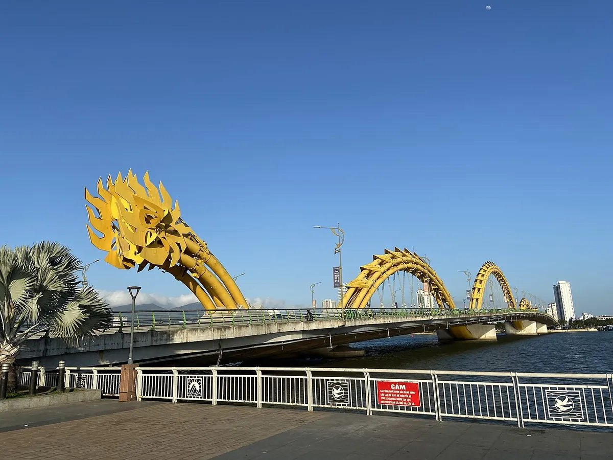 The Dragon Bridge by day, its golden arches rising over the road, seen from a riverside promenade