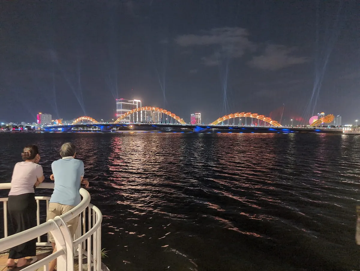 The Dragon Bridge lit in orange across the river at night, with spectators on a riverside railing in the foreground