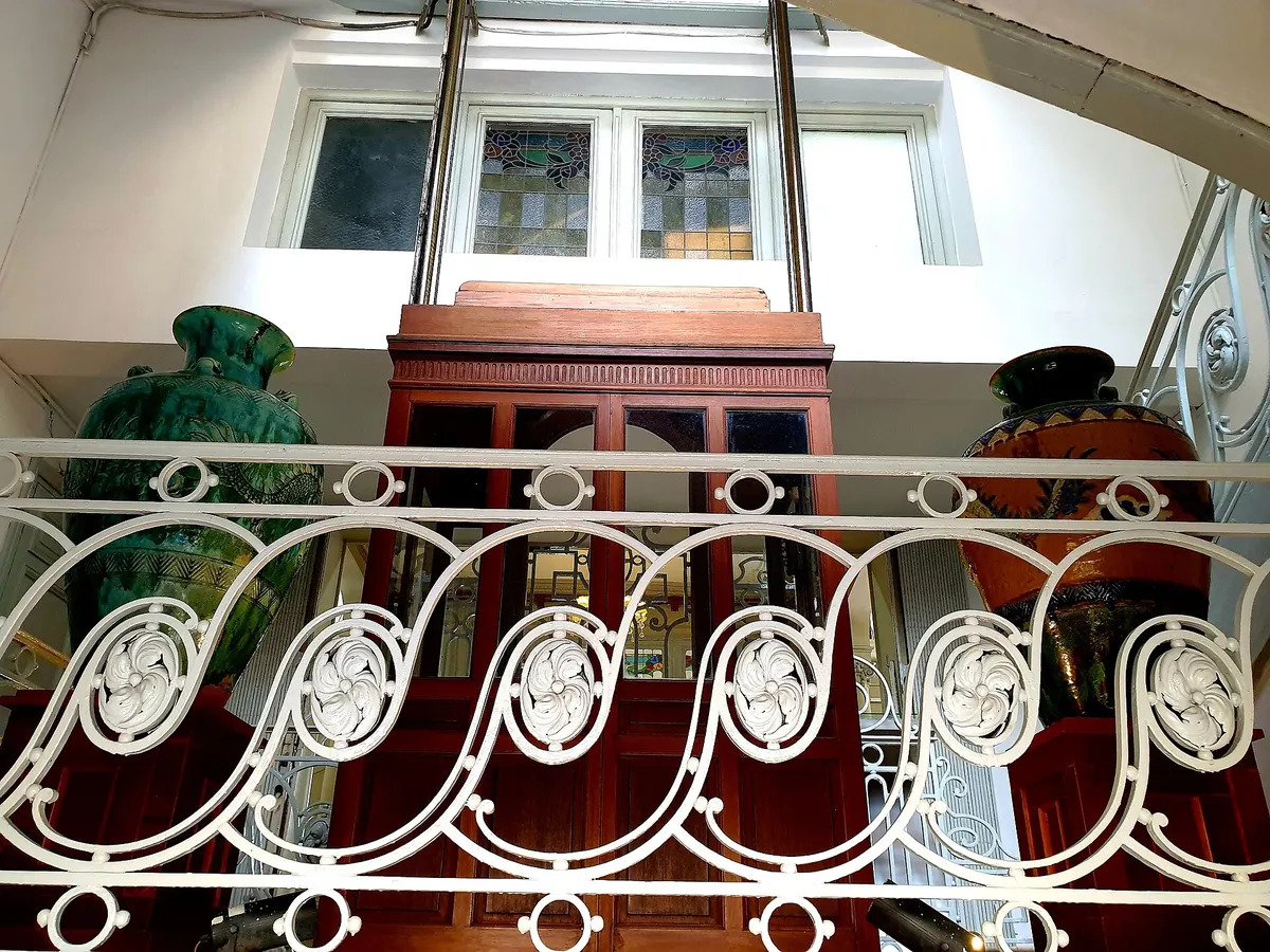 A white wrought-iron stair railing in front of a tall wooden cabinet and large glazed jars, under stained glass windows