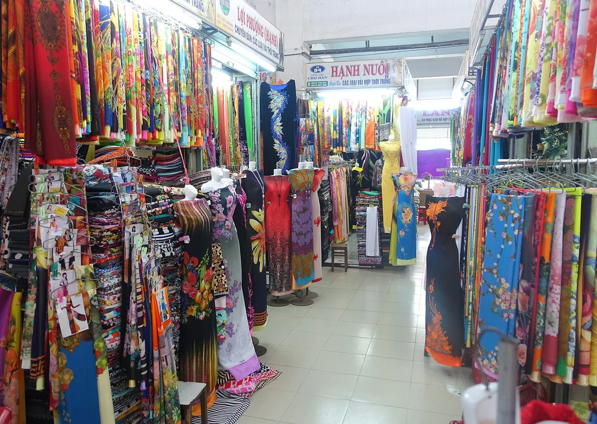 Bolts of fabric and hanging ao dai along a narrow aisle in Han Market