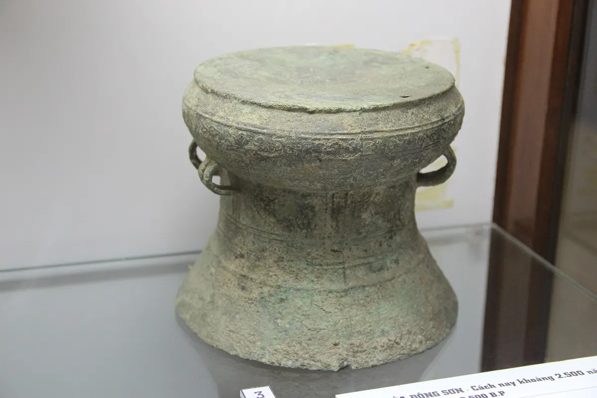 A small green bronze drum with two handles in a glass case