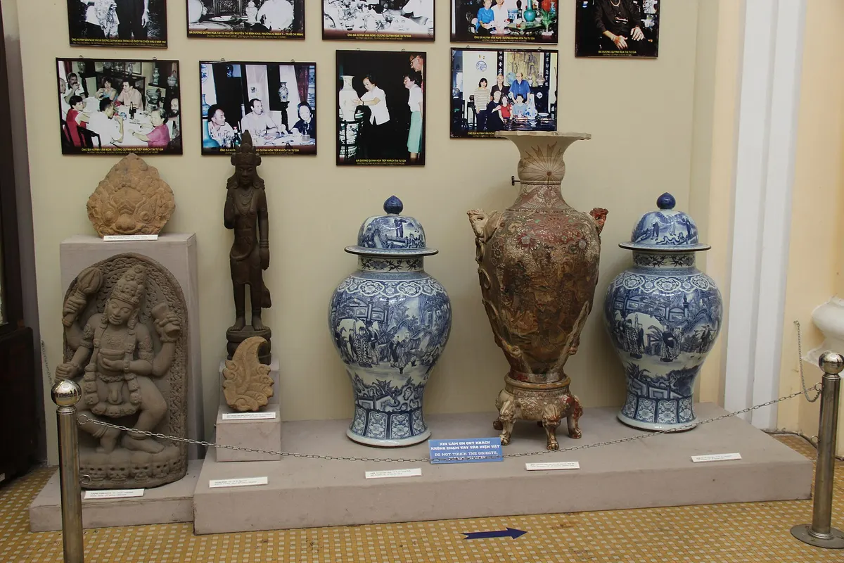 Stone and wooden statues beside large blue and white vases on a low platform, with photos on the wall behind