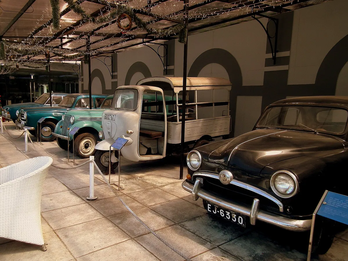 Old cars and a small bus parked in a row under a covered gallery