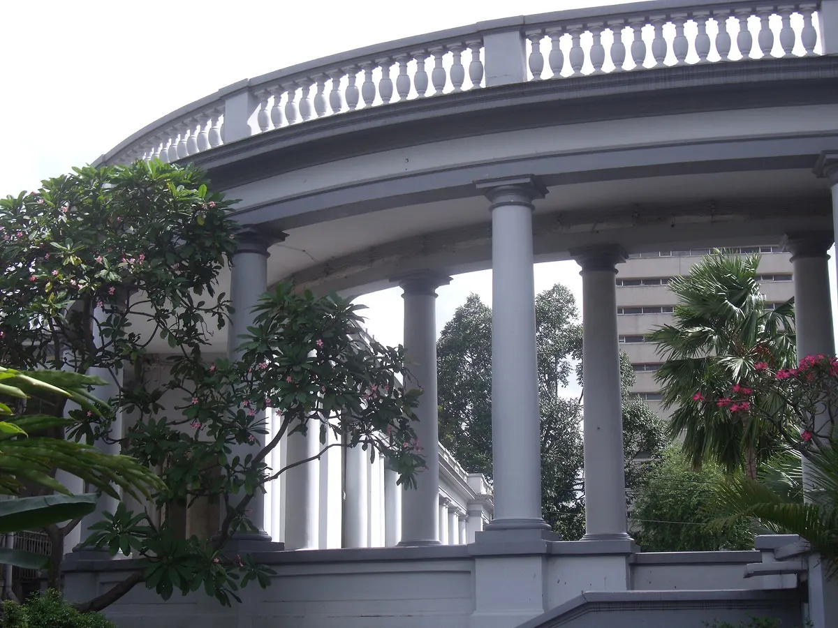 A curved colonnade with a balustrade along the top, frangipani trees in front