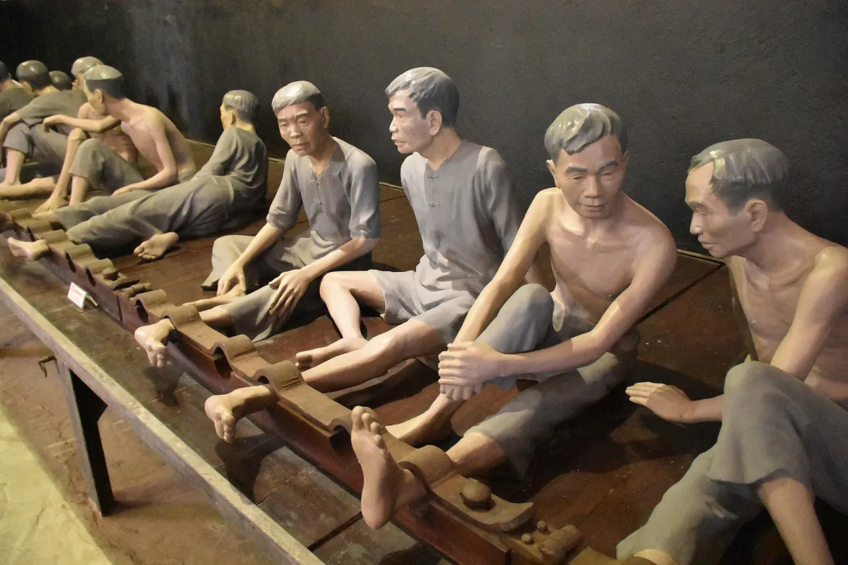 Life-size figures of prisoners sitting in a row with their ankles locked in wooden stocks