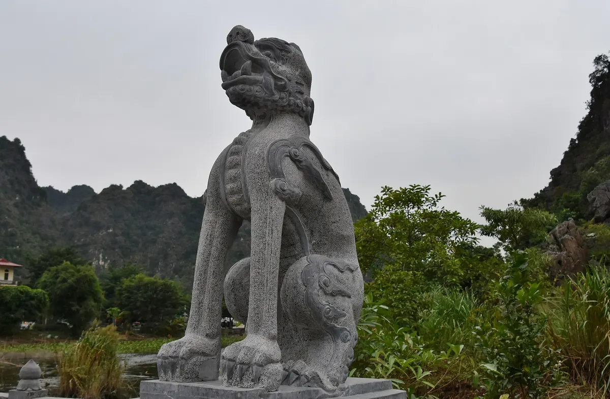 A grey stone guardian animal statue at Hoa Lu, with limestone hills and trees behind
