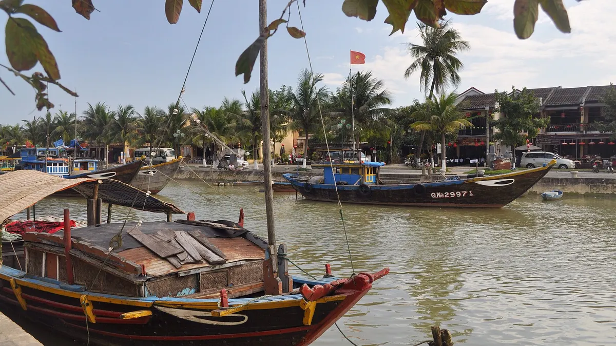 Small wooden boats moored on the river at Hoi An, with palm trees and old houses on the far bank