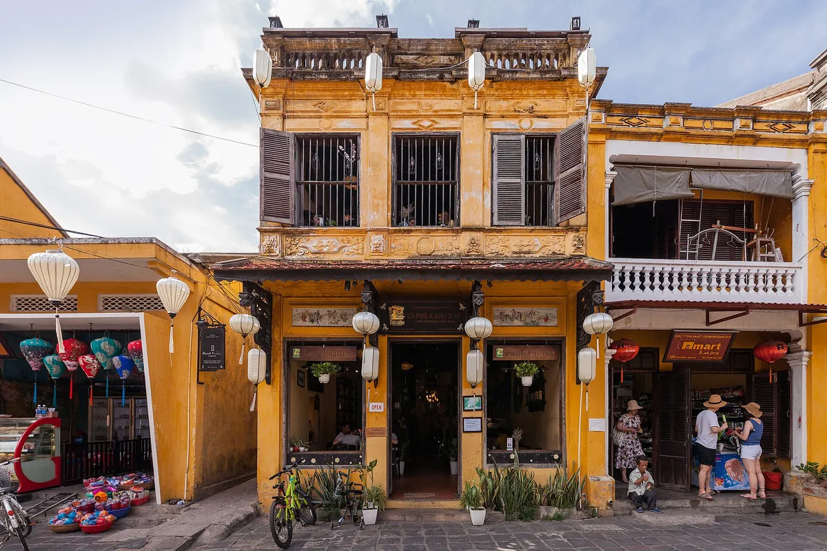 A two-storey yellow shophouse with wooden shutters and a balcony, lanterns hanging over the pavement