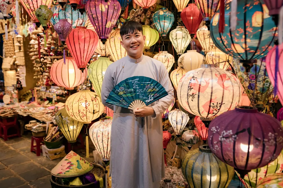 A smiling man in a pale grey ao dai holds a painted fan in a lantern shop crowded with silk lanterns