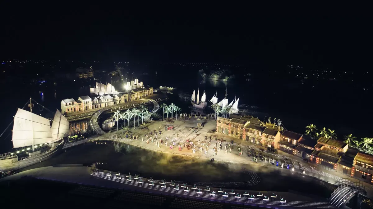 Aerial night view of the Hoi An Memories stage: a long lit stage with hundreds of performers, a row of shophouses, a red bridge and sailing ships along the river