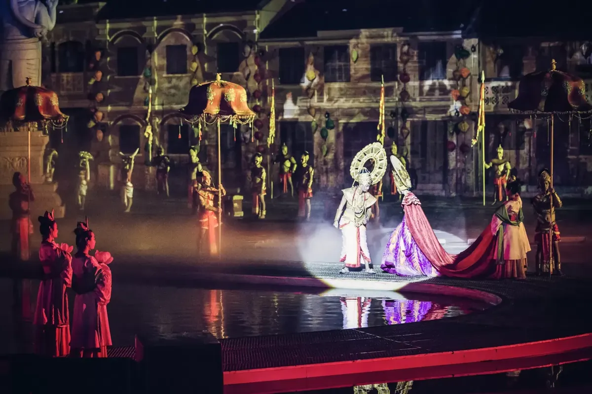 Performers in Cham-style costumes and tall headdresses on a lit stage with a reflecting pool, in front of a large painted palace facade