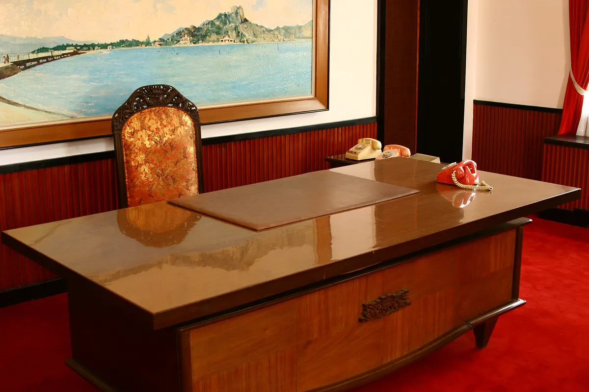 A polished wooden desk and high-backed chair in front of a painted seascape, with a red carpet on the floor