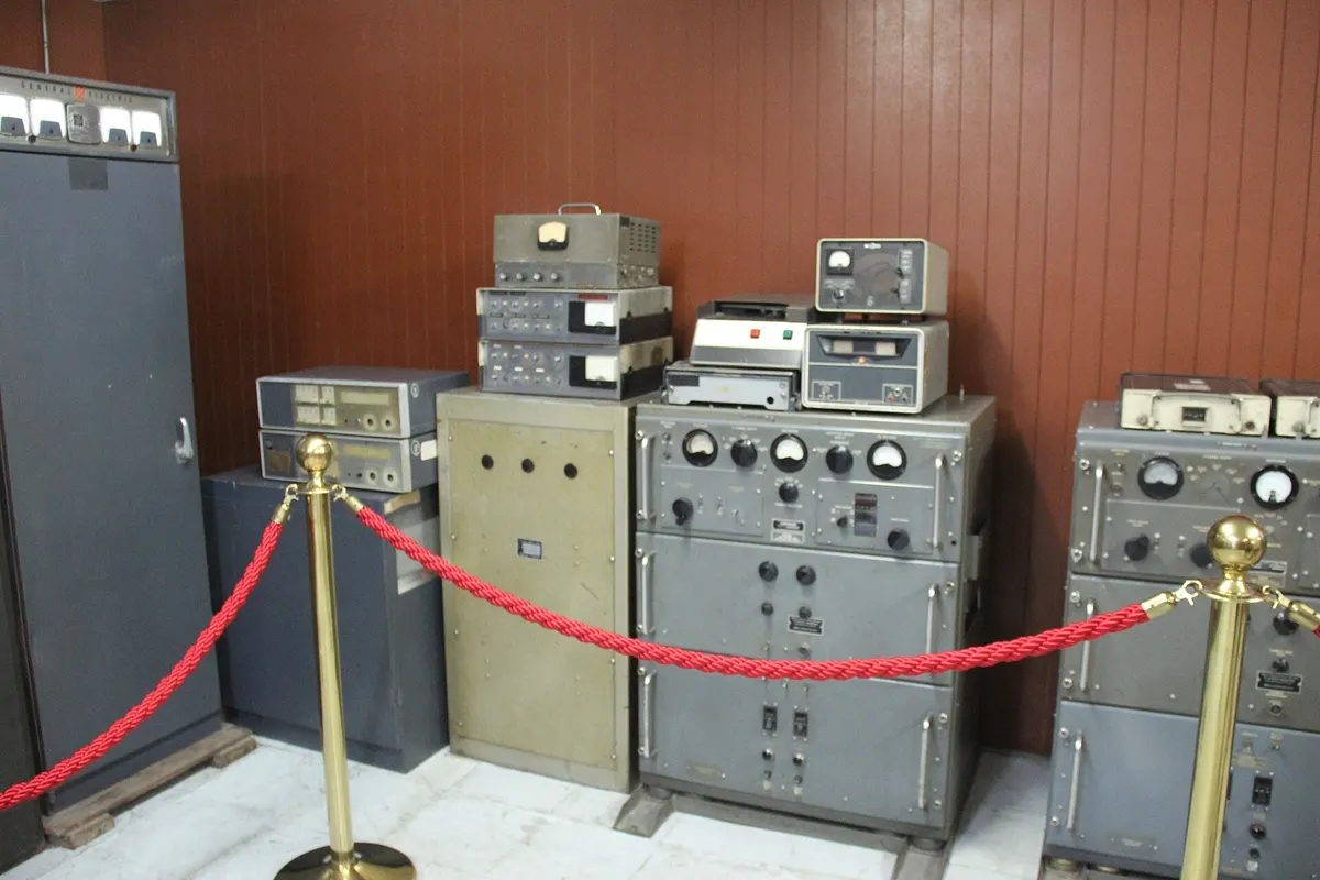 Grey radio transmitters and receivers lined up behind a red rope in a wood-panelled room