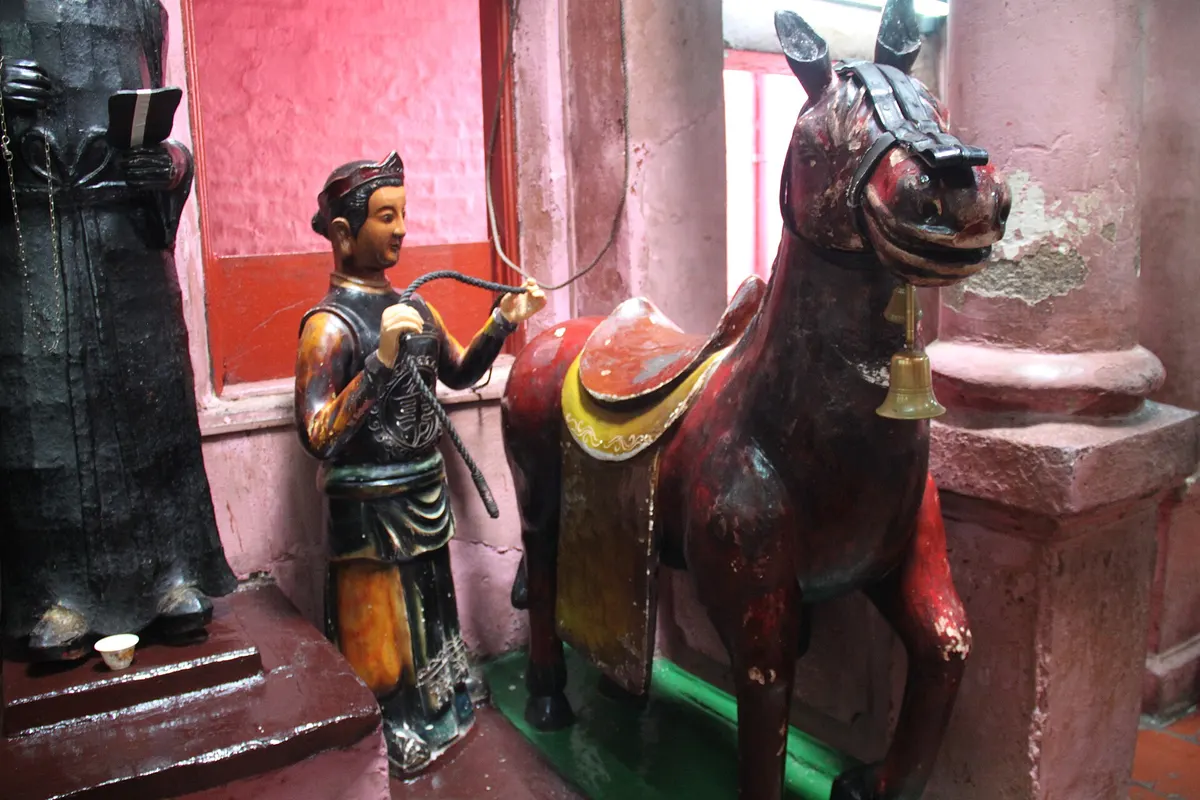 A painted wooden horse and a small figure of a groom holding its reins beside a pink pillar inside the temple