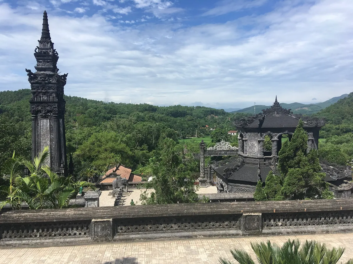 A dark stone pavilion and a tall pillar at the tomb, seen from a terrace with forested hills behind