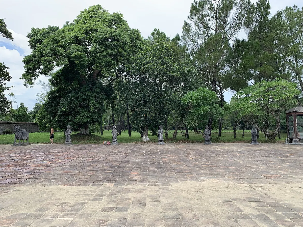 Stone mandarin statues standing in a row along the edge of the paved salutation court, with tall trees behind