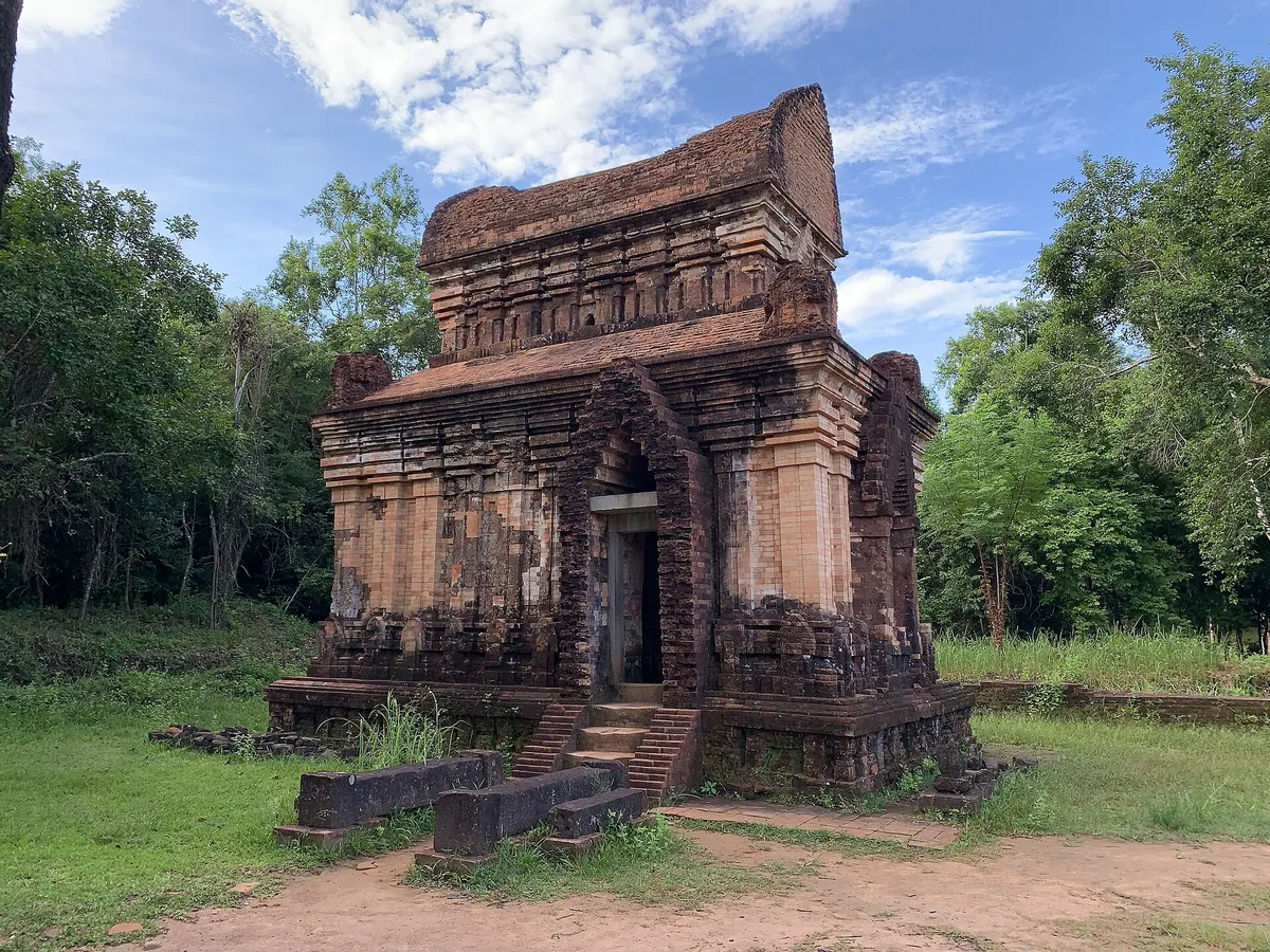 Temple E7 at My Son, a single brick tower with a carved doorway and stepped base, framed by trees
